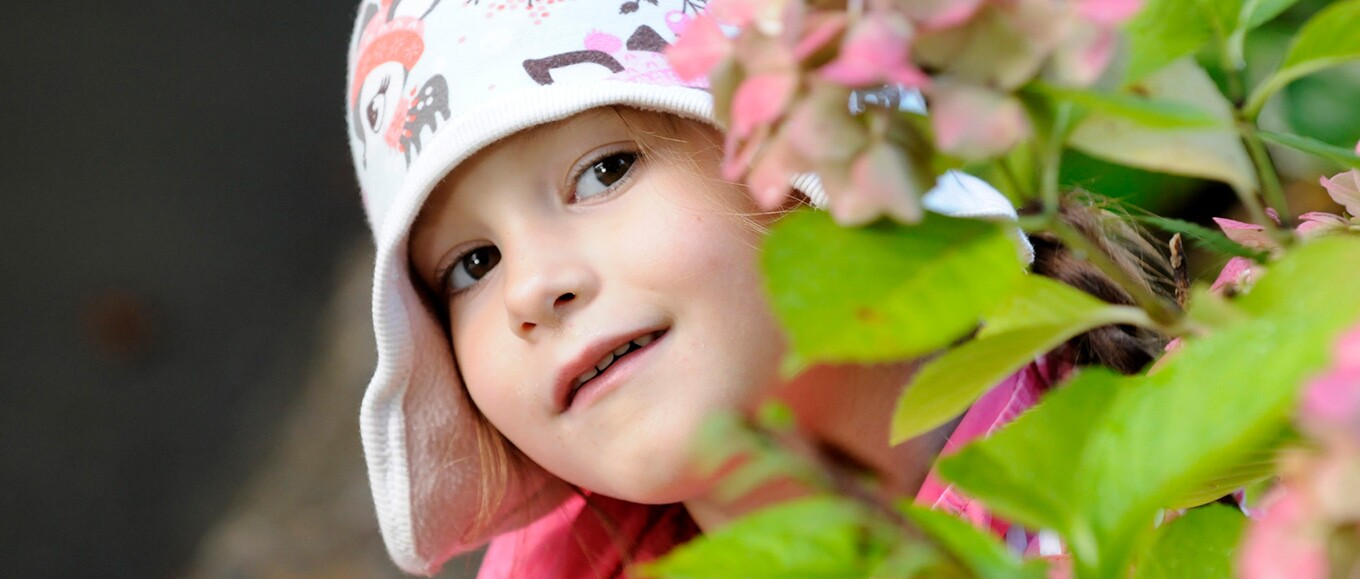 Ein Mädchen mit einer Tiermotiv-Mütze schaut hinter einer Blume hervor