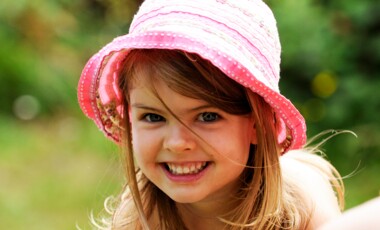 Ein Mädchen mit einem pinken Sommerhut und langen Haaren grinst in die Kamera