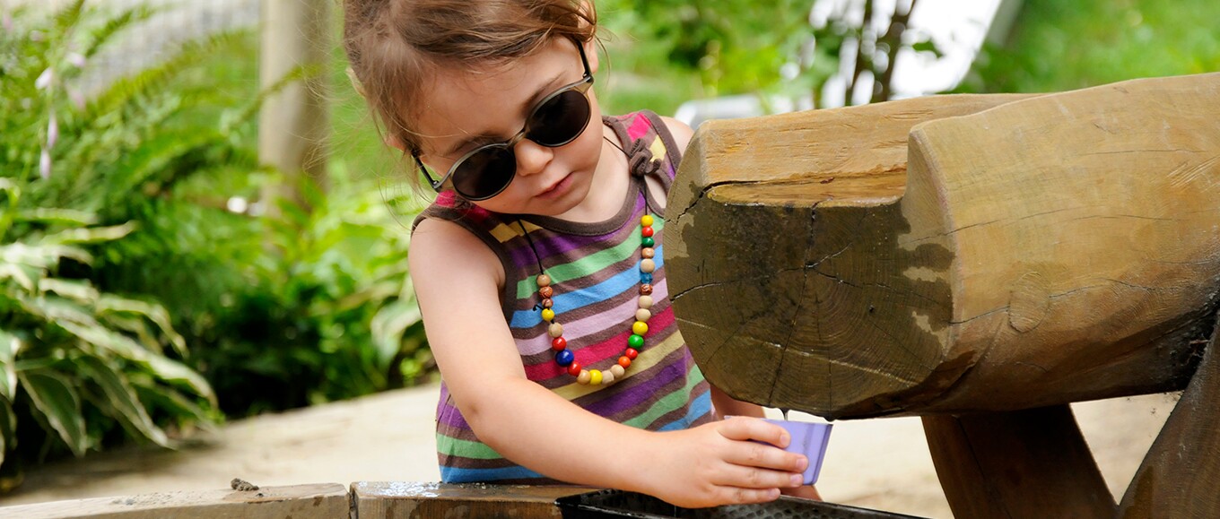 Ein bunt gekleidetes Kind mit Sonnenbrille spielt mit einem Becher am Wasserspielplatz